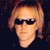 Pixelated close-up of a person with light brown hair and dark sunglasses against a dark background.