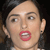 Pixelated close-up of a person with dark hair, light skin, and red lips, mouth open in a speaking expression against a dark background.