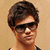 Pixelated close-up of a person with short dark hair, wearing black sunglasses and a white-and-black top against a beige background.