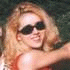 A woman with long blonde hair appears in two : first wearing dark sunglasses and a red top, then removing the sunglasses to reveal her eyes while holding a large black hat
