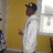 A pixelated figure in a white outfit and dark cap stands against a yellow wall with a window, holding an object near their waist.