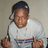 A pixelated image shows a person in a white shirt with a colorful stripe, wearing a dark cap and a necklace, making a gesture with both hands, next to blue with text