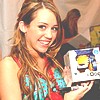 A woman with long brown hair smiles while holding a white box with a yellow and blue graphic on it, wearing a sleeveless multicolored top.