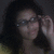 A person with dark curly hair and glasses adjusts their eyewear against a dark background, while adjacent text reads "OMG 90 SMEXXy" in yellow pixelated letters.
