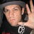 Pixelated image of a person wearing dark clothing and a gray cap, holding an object near their face with one hand raised.