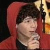 A person in a red hooded jacket holds a microphone, with a small red heart symbol visible in the background.