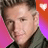 A person with styled hair wears a dark green collared shirt against a pink-to-purple gradient background, with a red square containing a white heart symbol in the top-right corner.
