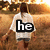 Pixelated image of a person in white shirt with black text 'he' on chest, standing against blurred brown and orange background.
