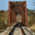 A pixelated image shows a rusted metal gate with a central archway, set against a blurred background of greenery and a light blue sky.