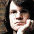 A person with dark hair rests their chin on a hand, wearing a black top and patterned sleeves against a textured background.