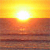 Pixelated sunset scene with bright yellow-orange sky transitioning to deep orange and brown water, centered white sunburst at horizon.