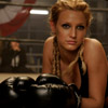 A woman with blonde braided hair wearing a black halter top and black boxing gloves, leaning forward with a serious expression in a gym setting.