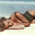 A pixelated image shows a person lying on their back on a light surface, wearing dark shorts and a dark top, with arms bent and hands near the head, against a gradient background.