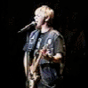 identical repeated images shows a person with short blonde hair wearing a dark shirt and holding a light-colored electric guitar on a dark stage with a microphone