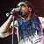 A person with long hair sings into a microphone, wearing a blue and white sleeveless shirt, with a dark background.