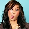 A person with long dark brown wavy hair and light skin extends their tongue out while wearing a brown jacket over a white top against a light blue background.