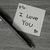 A pixelated grayscale icon shows a tilted white card with 'I Love You' text and a heart symbol, held by a black pen-like object against a gray background.