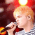 A person with short light brown hair sings into a microphone, wearing a striped shirt against a red background with blurred lights.
