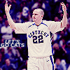 A basketball player in a white jersey with blue accents raises both arms, standing against a blurred crowd background.