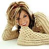 A woman with layered blonde hair lies on her stomach wearing a beige knitted sweater, resting her chin on her hands with a broad smile.
