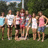 Group of people standing on grass with trees in background, some wearing white or pink tops and dark shorts.