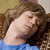 A pixelated close-up of a person with long brown hair wearing a blue top, resting their head on a dark surface against a brown and beige background.