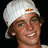Pixelated image of a person wearing white cap with red logo, smiling against black background.