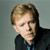 Pixelated portrait of a person with light brown hair wearing a dark jacket over a blue shirt against a muted gray background.