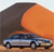 A pixelated image shows a light blue sedan with dark windows parked on a brown surface under a large orange circular shape and a dark angular structure.