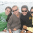 Four people pose together outdoors with greenery and a light sky background; two wear dark sunglasses, one has short brown hair, another wears a black shirt.