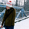 A person wearing a white knit hat with red patterns and a red pom-pom stands on a snowy bridge with blue metal railings, trees in the background.