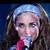 A woman with long brown hair and a light blue headband sings into a red microphone against a dark background.