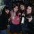 Group of people in dark clothing with one person holding a silver and red object, seated on a brown surface with a blurred background.