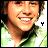 Pixelated close-up of a smiling person with light brown hair wearing a green and white garment, set against a blurred background with a small black symbol on the right.