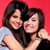 Two women embrace against a red background, one in light gray shirt and the other in black top with green wristband.