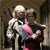 Two figures in a pixelated image: one in white with a pink chest area, the other in dark clothing holding a red object, standing close together against a brown background.