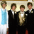 Four people stand in a row; one wears a light blue dress, three wear black tuxedos with white shirts and bow ties, one has a white pocket square.