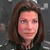 Pixelated portrait of a person with shoulder-length brown hair against a dark gray background, wearing a dark top.