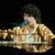 A person with short dark hair wearing a gold patterned jacket over a white top, positioned behind a reflective surface.