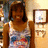 A person in a sleeveless top with blue and red patterns stands against a wall with framed pictures, smiling at the camera.