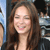 A static image of a person with long brown hair smiling, wearing a dark top against a blurred background with blue and gray tones.