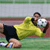 Pixelated static image of a soccer player in yellow jersey diving to save a ball on a field with green and red background stripes.