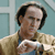 A pixelated image shows a person with dark hair wearing a light brown jacket over a blue shirt, holding a white object against a light blue background.