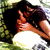 Two figures in a close embrace, one with dark hair and glasses resting on the other's shoulder against green foliage background.