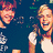 Two pixelated figures with light hair against a dark blue background; one figure on the right points toward the viewer while both display broad smiles.