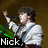 A person in a white shirt with blue straps holds a microphone, standing before green stage lights and a brown background.