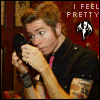 A person with light brown hair holds a black microphone near their mouth, wearing a black shirt with a pink undershirt visible, tattoos on their arm, and a black wristband, with text "I FEEL PRETTY" and a white symbol on a red background.