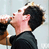 A person with dark spiky hair sings into a microphone, wearing a black shirt and small ear piercing; text overlays change