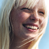 A close-up of a person with long blonde hair smiling broadly, showing teeth, against a light blue background.