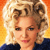 Pixelated portrait of a person with voluminous blonde hair styled in large curls, wearing a dark blue top against a gradient orange-yellow background.